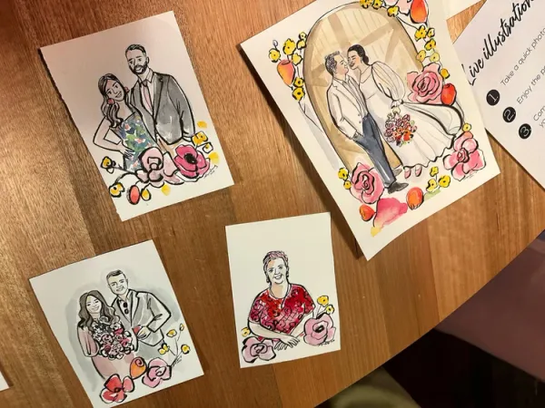 Ink and watercolour wedding guest portraits framed with pink and yellow flowers on a wooden table, by Alyssa Bermudez Art