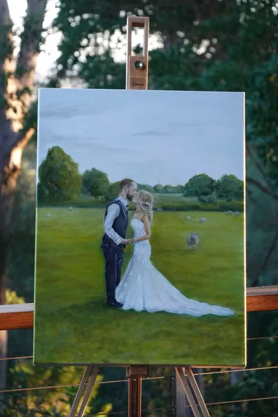 Oil painting on an easel of a bride and groom kissing in a green paddock with grazing sheep, by Darcy Hart