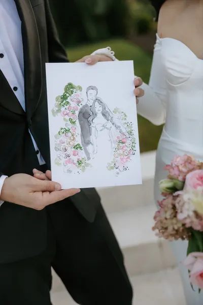 Couple holding a live watercolour and ink illustration of themselves framed by pink roses, by Grace Choi Artist