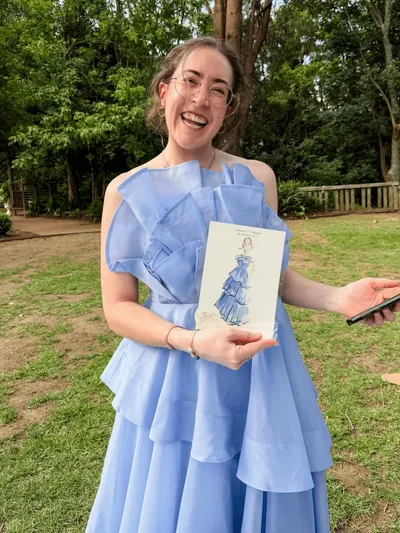 Smiling wedding guest in a blue ruffled gown holding her live watercolour fashion portrait, by Graced Goods